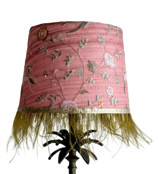 Pink lampshade with floral patterns and gold fringe on a plain background