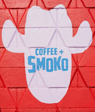 White cactus-shaped sign with 'Coffee + Smoko' text on a red brick wall.